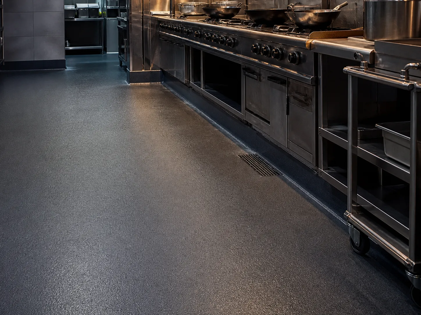 Dark grey textured resin floor beside a commercial cooking line, stainless drain and trolley wheel